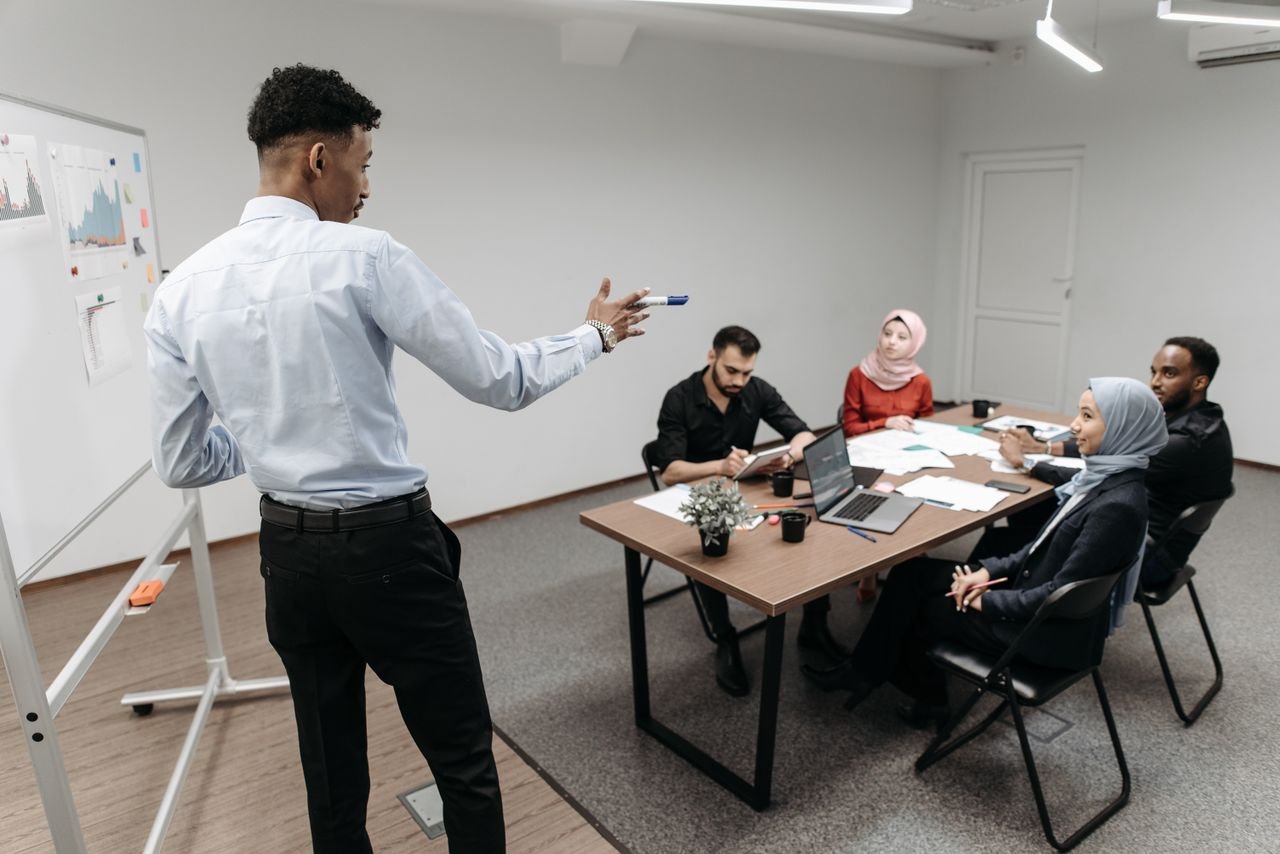 A multicultural business team engaged in a discussion during an office meeting with a whiteboard and laptops.