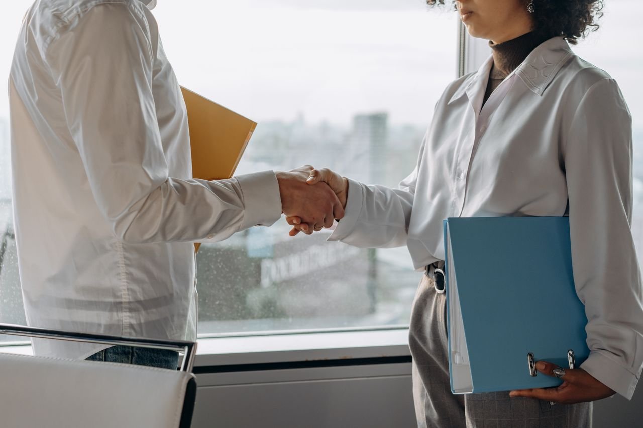 Two professionals in business attire shaking hands in an office setting with view.